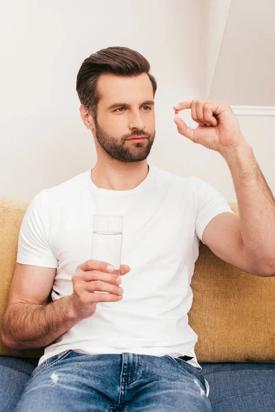 Hombre guapo sosteniendo un vaso de agua y mirando la píldora en el sofá - foto de stock