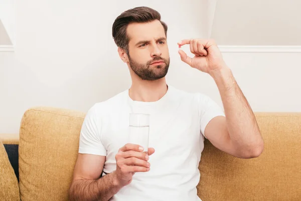 Hombre reflexivo sosteniendo píldora y vaso de agua en el sofá en casa — Stock Photo