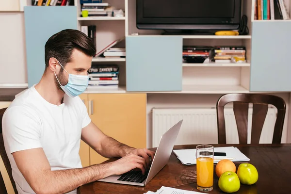Vista laterale dell'uomo in maschera medica che lavora sul computer portatile vicino a succo d'arancia e frutta sul tavolo — Foto stock