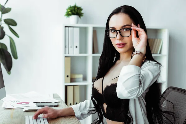 Secrétaire sensuel en soutien-gorge et lunettes regardant la caméra et l'utilisation de l'ordinateur dans le bureau — Photo de stock