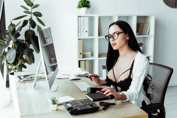 Secretaria sensual usando teléfono mientras trabaja cerca de computadora y papeles en la oficina - foto de stock