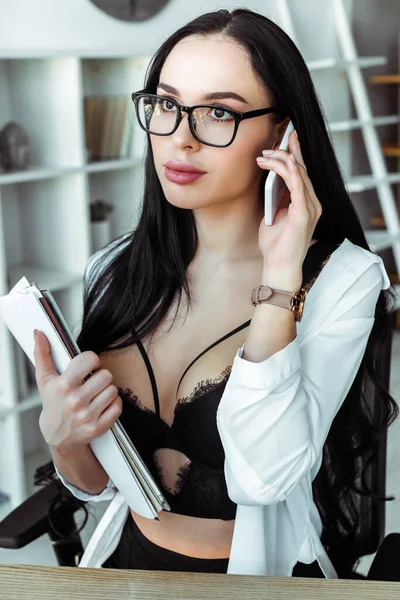 Attractive secretary in eyeglasses and bra talking on smartphone and holding papers in office — Stock Photo