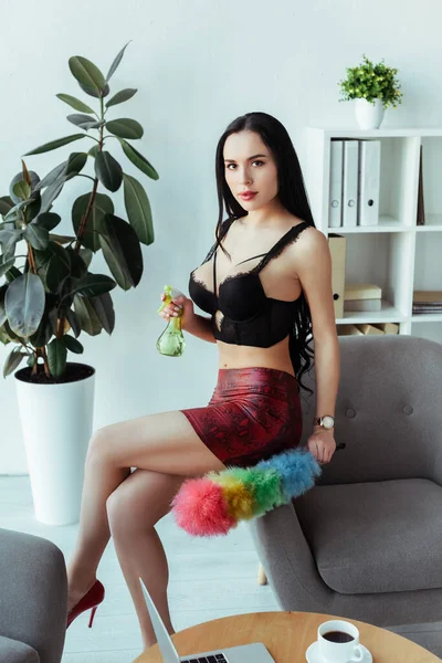 Selective focus of seductive secretary holding spray bottle and dust brush on armchair in office — Stock Photo