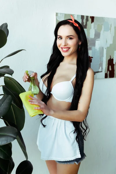 Sensual chica sonriente sosteniendo botella de spray y trapo cerca de la planta en casa - foto de stock