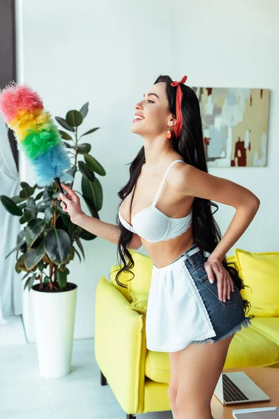 Side view of attractive girl in bra and apron smiling and holding dust brush in living room — Stock Photo