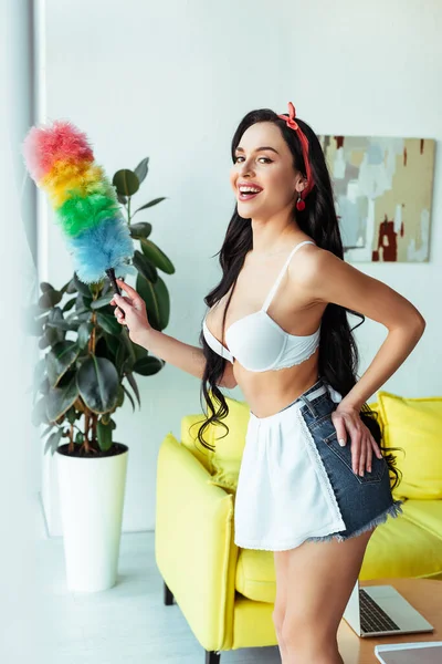 Side view of seductive woman in bra and apron holding dust brush and smiling at camera at home — Stock Photo