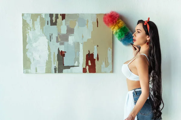 Side view of sexy girl in bra and apron cleaning painting with dust brush at home — Stock Photo