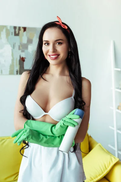 Hermosa chica en sujetador y delantal sonriendo a la cámara mientras sostiene el ambientador en casa - foto de stock