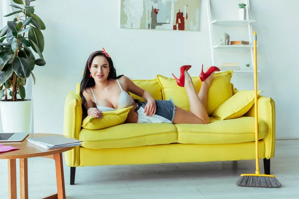 Sensual smiling woman looking at camera while lying on couch near broom — Stock Photo