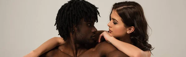 Sexy interracial pareja abrazándose y mirando uno al otro aislado en gris, cultivo panorámico - foto de stock