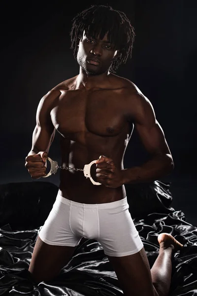 Sexy homme afro-américain en boxeurs blancs tenant les menottes sur le lit sur noir — Photo de stock