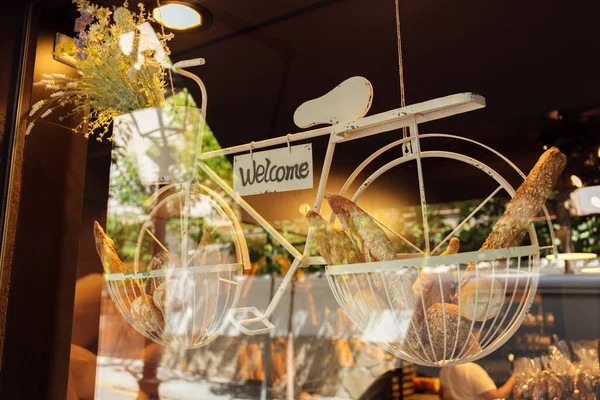 Dekoratives Fahrrad mit Blumen, Gebäck und Karte mit Willkommensschrift auf der Vitrine der Bäckerei — Stockfoto