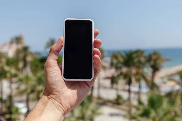 Cropped view of man holding smartphone with palm trees and sea coast at background — Stock Photo