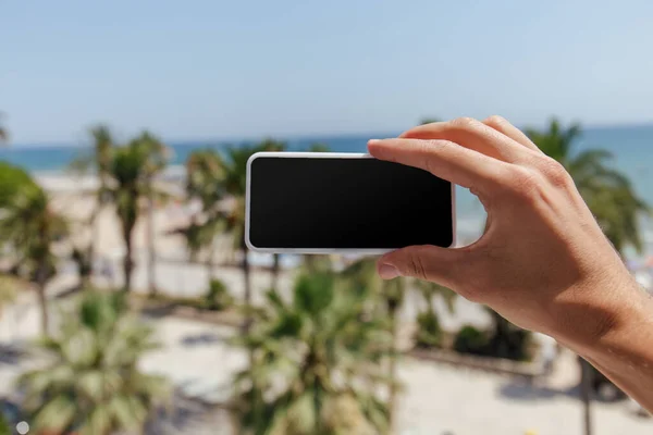 Cropped view of man holding smartphone with blank screen with sea coast at background — Stock Photo