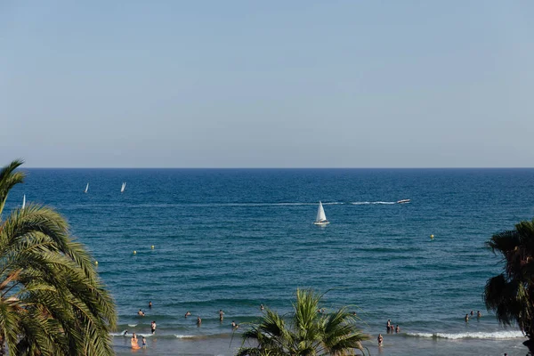 CATALONIA, ESPAÑA - 30 DE ABRIL DE 2020: Personas descansando en la playa con palmeras y yates en el mar al fondo - foto de stock