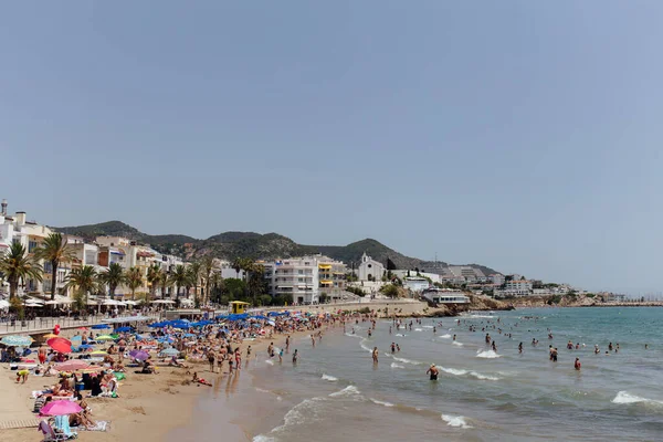 CATALONIA, ESPAÑA - 30 DE ABRIL DE 2020: Personas descansando en la playa de arena y nadando en el mar cerca de edificios y palmeras en la costa - foto de stock