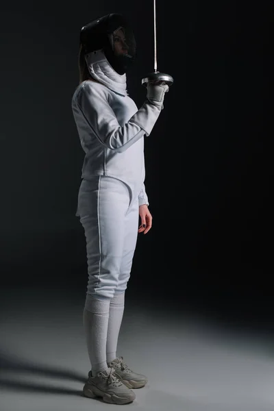 Side view of fencer in fencing suit and mask holding rapier on grey surface on black background — Stock Photo