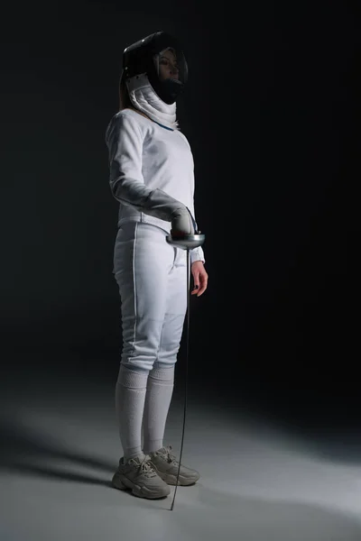 Fencer in fencing suit and mask holding rapier on grey surface on black background — Stock Photo