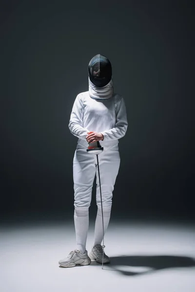 Fencer in fencing mask and suit holding rapier under spotlight on black background — Stock Photo