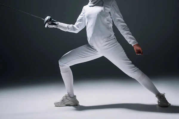 Cropped view of fencer in fencing suit training with rapier on white surface on black background — Stock Photo
