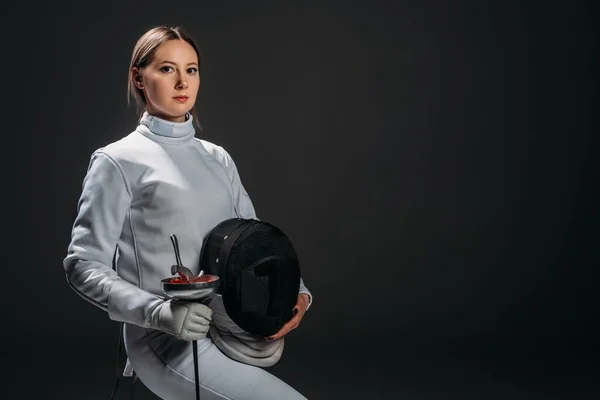 Beautiful swordswoman looking at camera while holding fencing mask and rapier isolated on black — Stock Photo