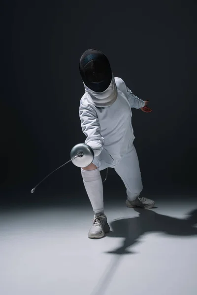 Esgrima em esgrima máscara segurando rapier durante o treinamento na superfície branca no fundo preto — Fotografia de Stock