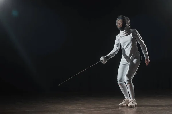 Swordswoman in fencing mask and suit training with rapier under spotlight on black background — Stock Photo