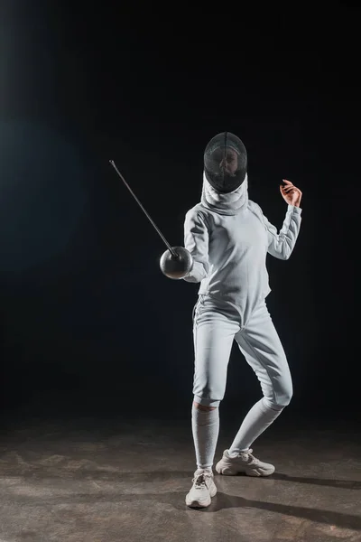 Fencer in fencing mask holding rapier while training under spotlight on black background — Stock Photo