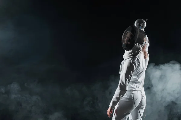 Rückenansicht der Schwertkämpferin mit Fechtmaske, die auf schwarzem Hintergrund mit Rauch in die Kamera blickt — Stockfoto