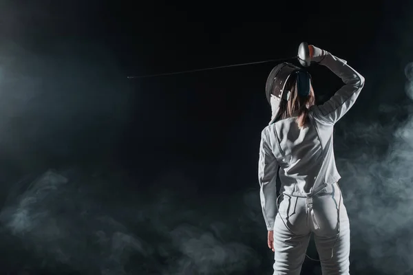 Back view of fencer holding rapier above head on black background with smoke — Stock Photo