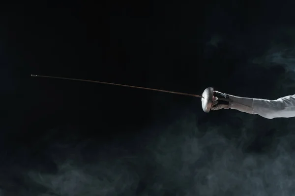 Cropped view of fencer in glove holding rapier on black background with smoke — Stock Photo