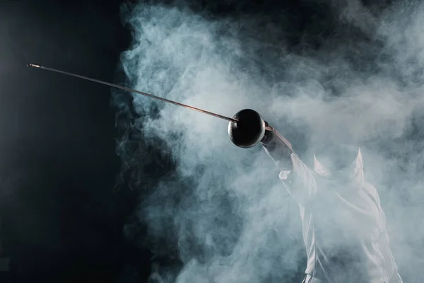 Low angle view of fencer holding rapier on black background with smoke — Stock Photo