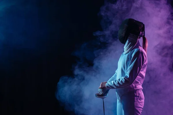 Side view of fencer in fencing mask holding rapier on black background with smoke — Stock Photo