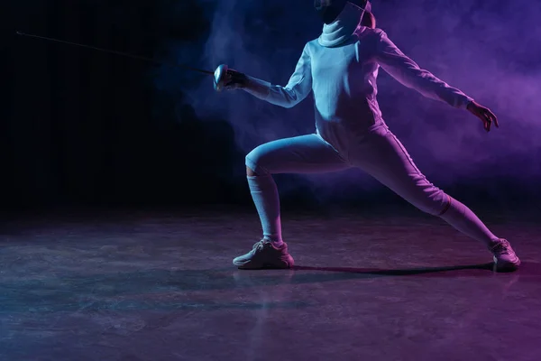 Cropped view of swordswoman fencing on black background with smoke and lighting — Stock Photo