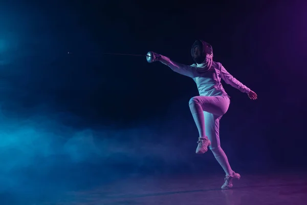 Esgrima en máscara de esgrima y entrenamiento de traje con estoque sobre fondo negro con humo e iluminación - foto de stock