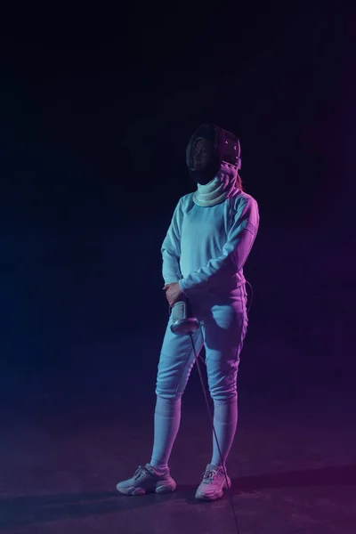 Swordswoman in fencing mask and suit holding rapier and looking away on black background — Stock Photo