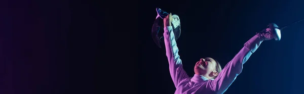 Excited and cheerful fencer holding rapier and fencing mask isolated on black, horizontal crop — Stock Photo