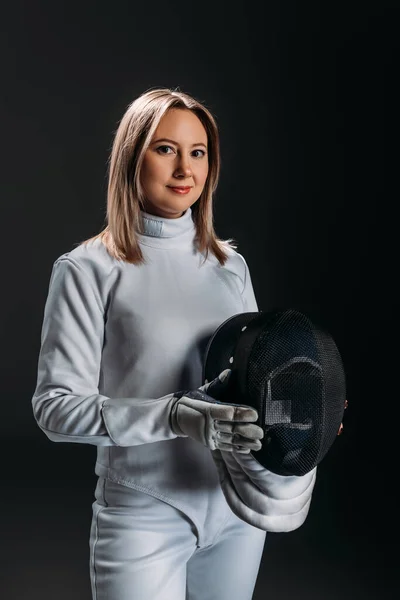 Attractive fencer in fencing suit holding mask and looking at camera isolated on black — Stock Photo