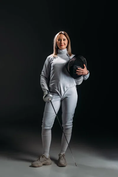 Beautiful fencer looking at camera while holding rapier and fencing mask on grey surface on black background — Stock Photo