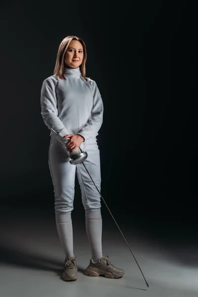 Beautiful fencer in fencing suit holding rapier on grey surface on black background — Stock Photo