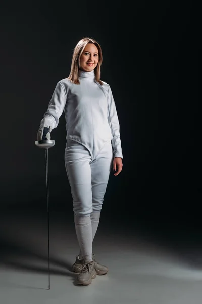 Smiling fencer in fencing suit and glove looking at camera while holding rapier on grey surface on black background — Stock Photo