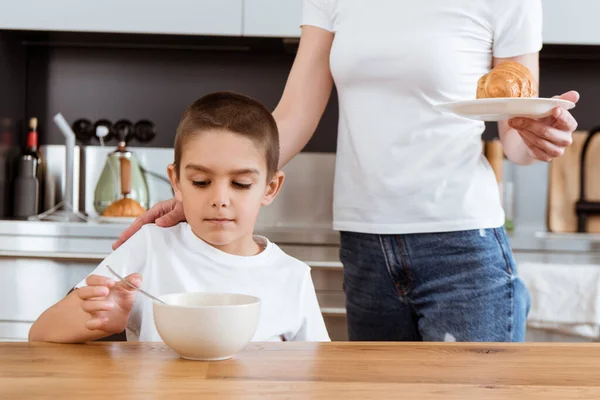 Mãe segurando croissant perto do filho tomando café da manhã na cozinha — Fotografia de Stock