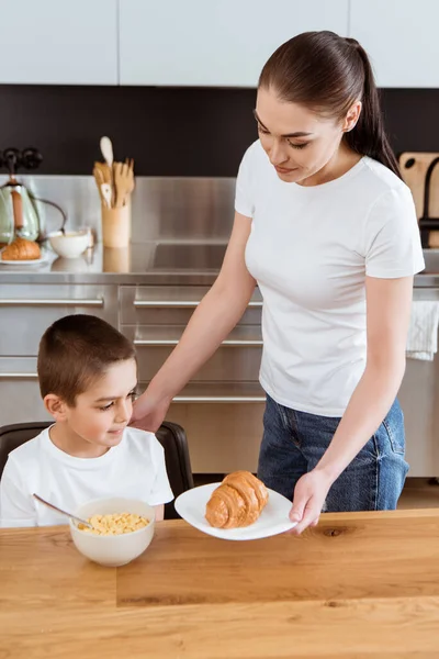 Mother holding croissant near son and bowl of cereals on kitchen table — Stock Photo