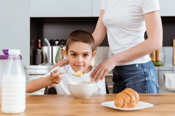 Foco seletivo do menino olhando para a câmera enquanto come cereais perto da mãe na cozinha — Fotografia de Stock