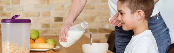 Orientação panorâmica da mãe derramando leite em cereais perto de filho sorridente à mesa na cozinha — Fotografia de Stock