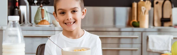 Panoramic crop of smiling kid looking at camera near cereals and milk in kitchen — Stock Photo