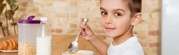 Cultivo horizontal del niño mirando la cámara cerca de los cereales y la leche en la mesa de la cocina - foto de stock