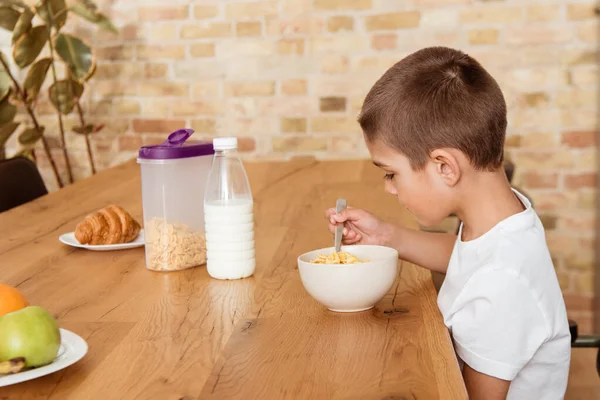 Side view of boy eating cereals near milk, croissant and fruits on table in kitchen — Stock Photo