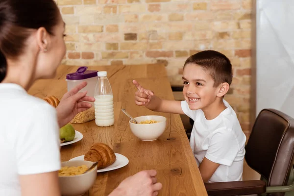 Selective focus of cheerful child pointing with finger at mother during breakfast in kitchen — Stock Photo