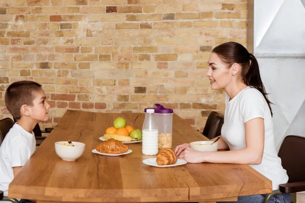 Vista lateral da mãe sorridente olhando para o filho perto de cereais e croissants na mesa da cozinha — Fotografia de Stock